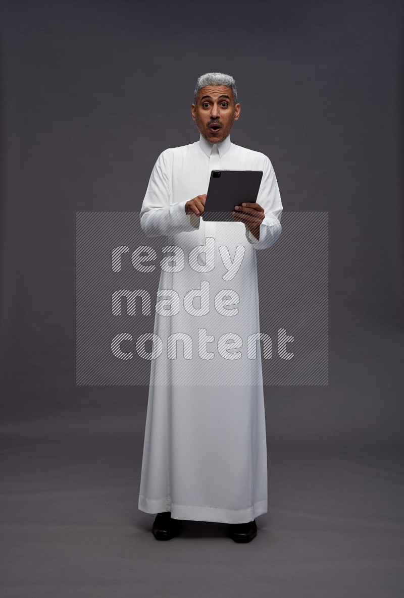 Saudi man wearing thob standing working on tablet on gray background