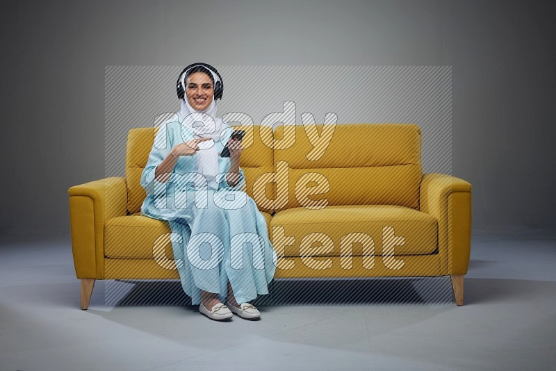 A Saudi woman wearing a light blue Abaya and a white head scarf sitting on a yellow sofa and holding an electronic while wearing headphone eye level on a grey background