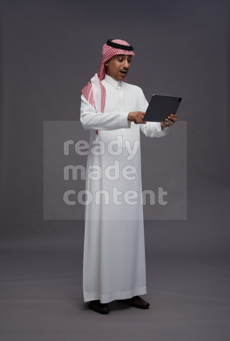 Saudi man wearing thob and shomag standing working on tablet on gray background