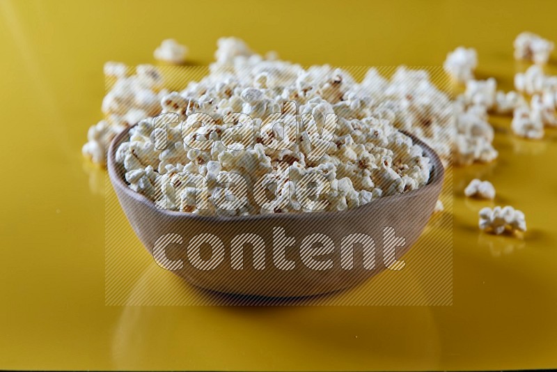 A brown pottery bowl full of popcorn with popcorn beside it on a yellow background in different angles