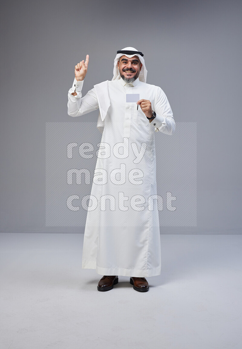 Saudi man Wearing Thob and white Shomag standing holding ATM card on Gray background
