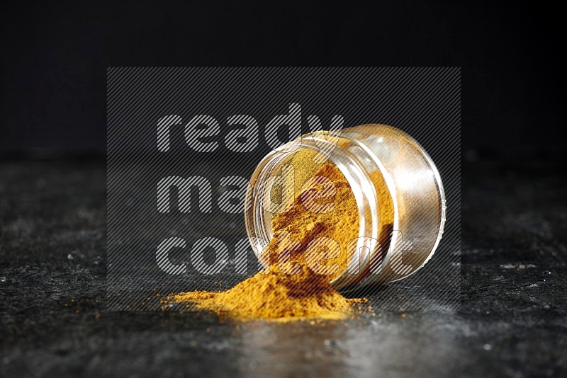 A flipped glass jar full of turmeric powder and powder spilled out of it on a textured black flooring
