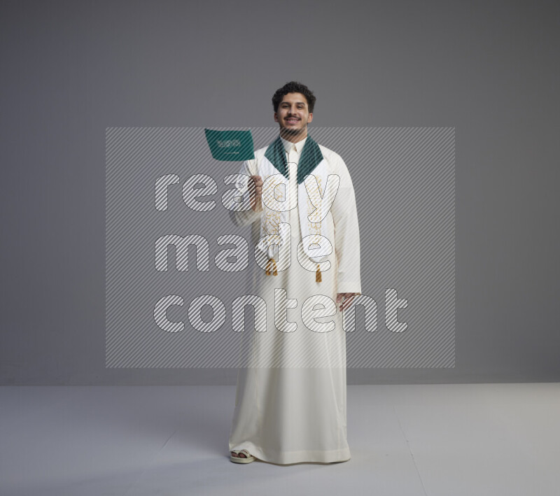 A Saudi man standing wearing thob and saudi flag scarf and holding small saudi flag on gray background