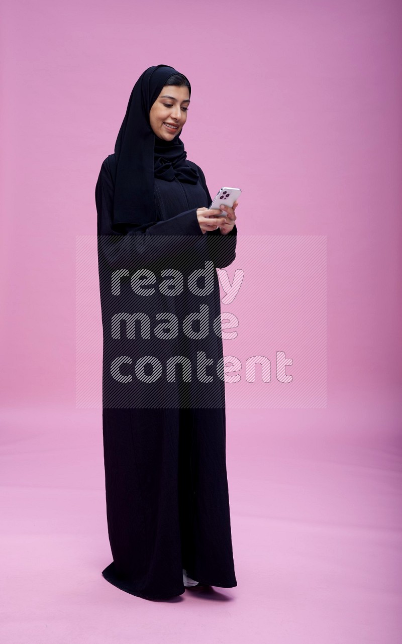 Saudi woman wearing Abaya standing texting on phone on pink background