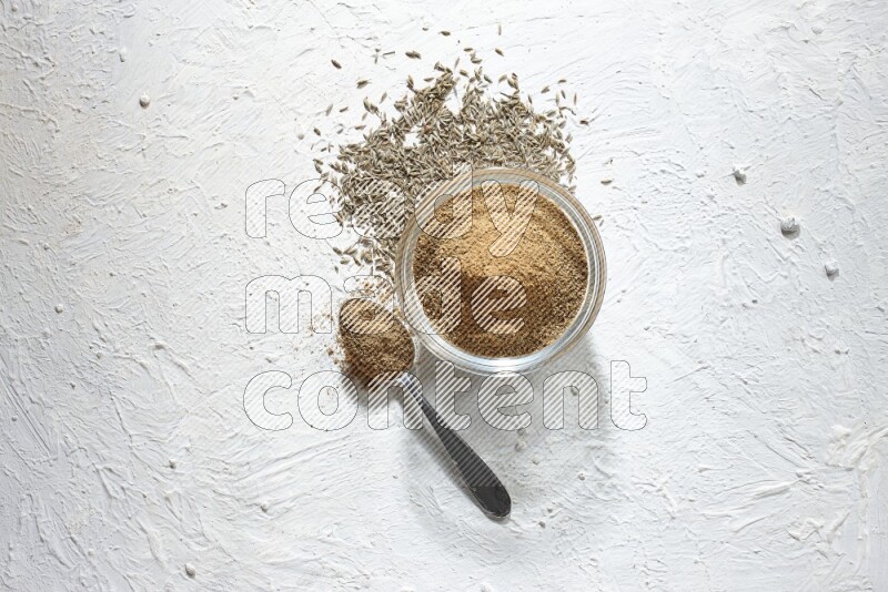 A glass bowl and metal spoon full of cumin powder and cumin seeds underneath it on textured white flooring