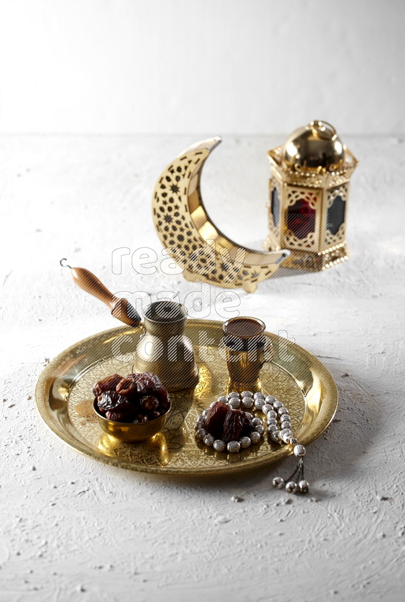 Dates in a metal bowl with coffee and prayer beads on a tray beside lanterns in a light setup
