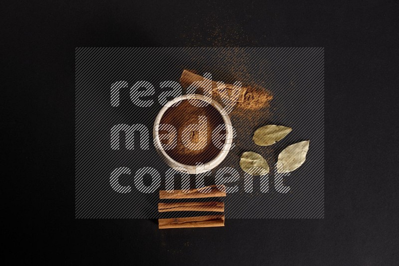 Cinnamon powder in a white pottery bowl and cinnamon sticks and laurel leaves on black background