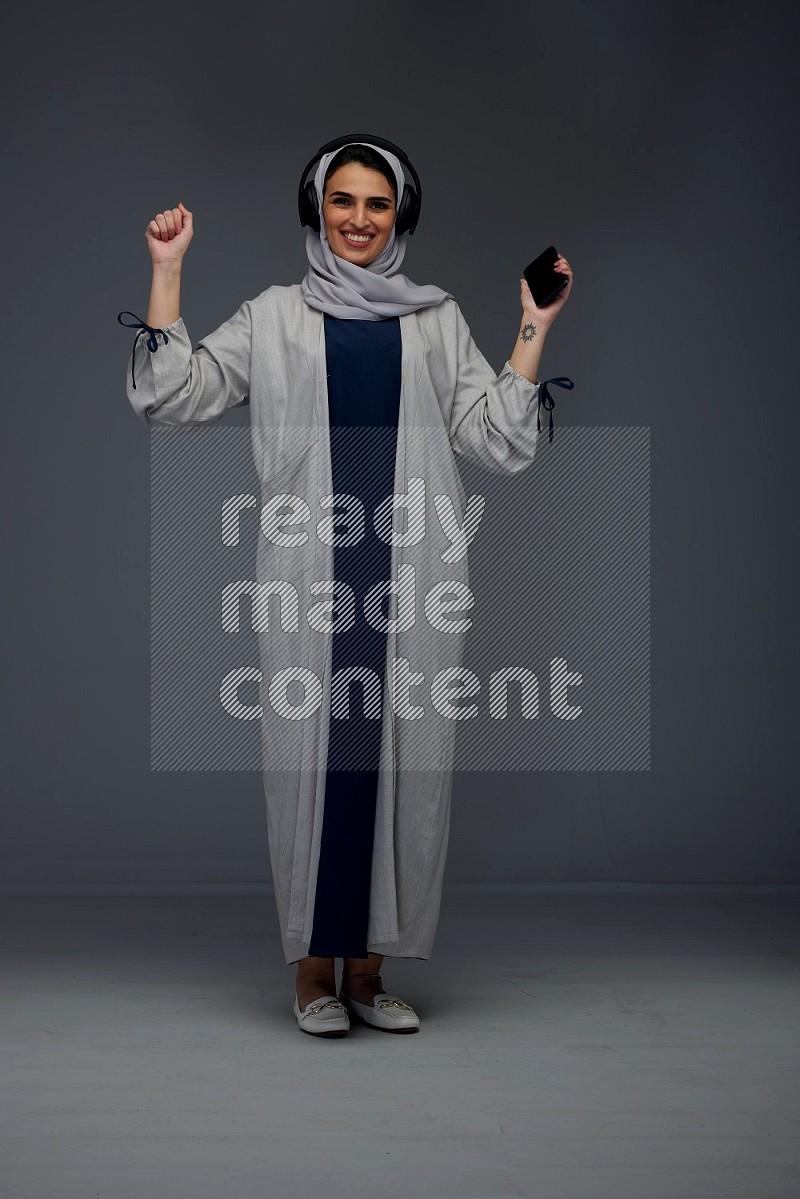 A Saudi woman wearing a light gray Abaya and head scarf standing and listening to music on a grey background