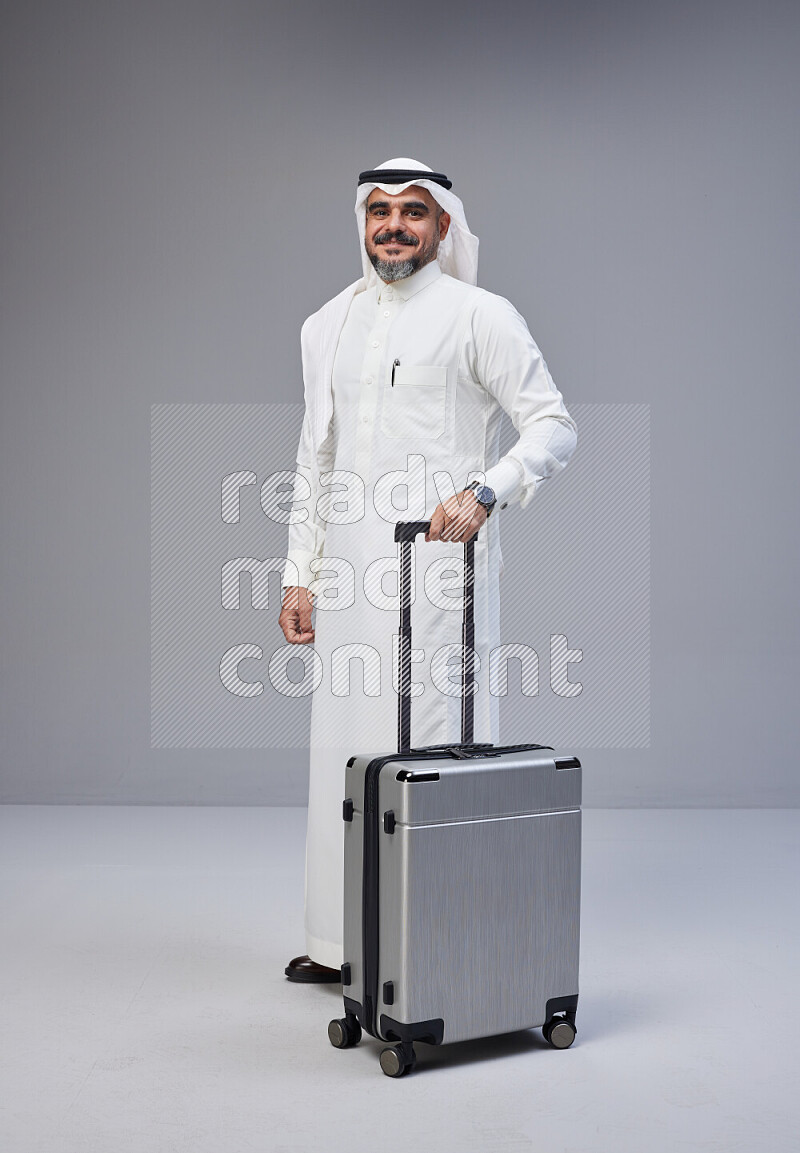 Saudi man wearing Thob and white Shomag standing holding Travel bag on Gray background
