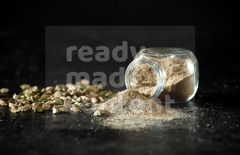A flipped glass spice jar full of cardamom powder and powder spilled out of it with cardamom seeds spreaded on textured black flooring
