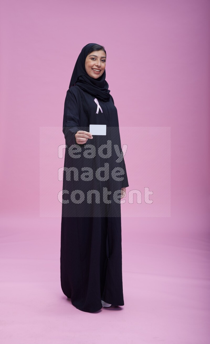 Saudi woman wearing pink ribbon on Abaya standing holding ATM card on pink background