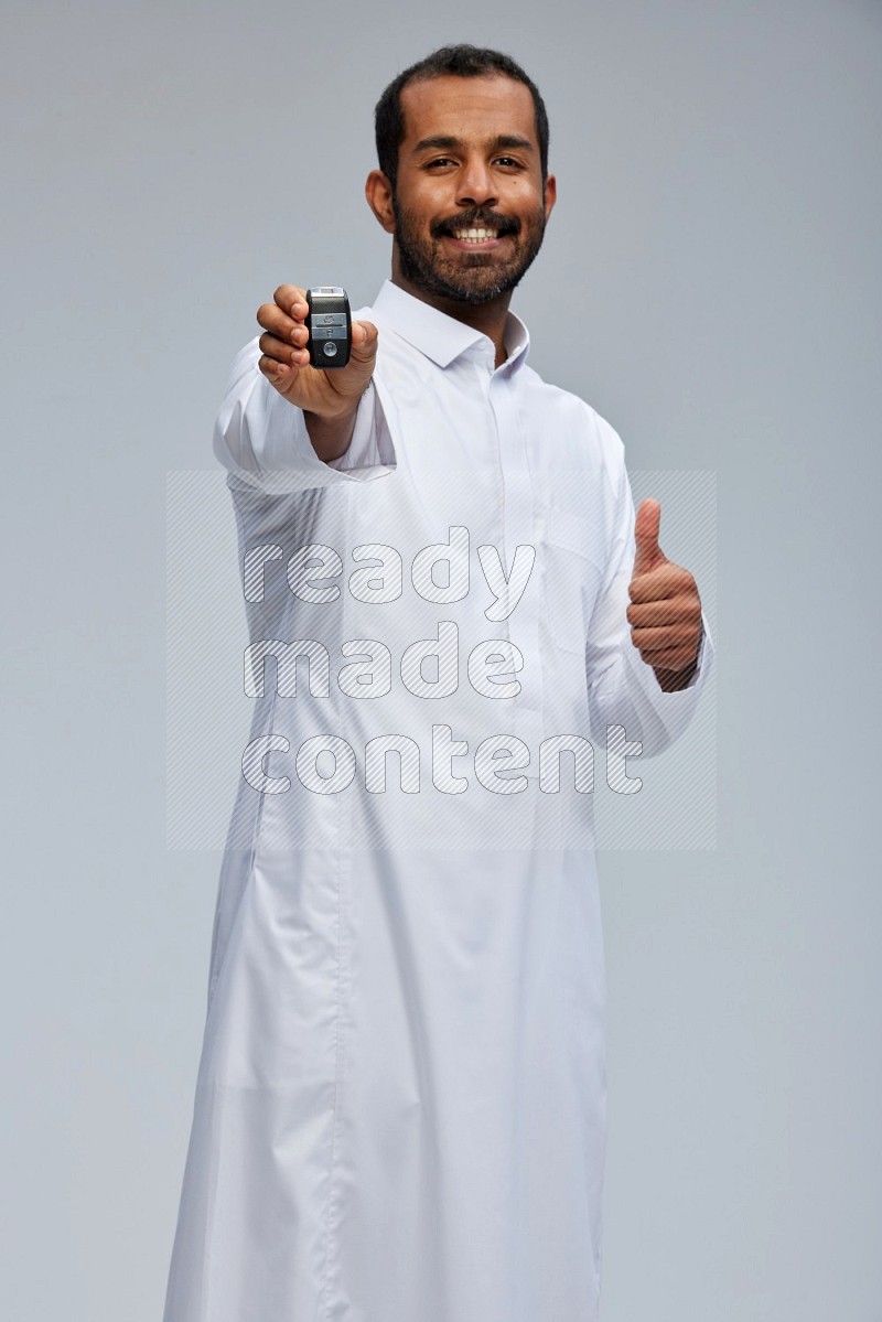 Saudi man wearing thob and shomag standing holding car key on gray background