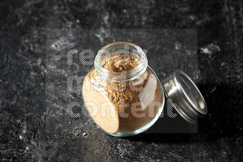 A glass spice jar full of allspice powder on a textured black flooring