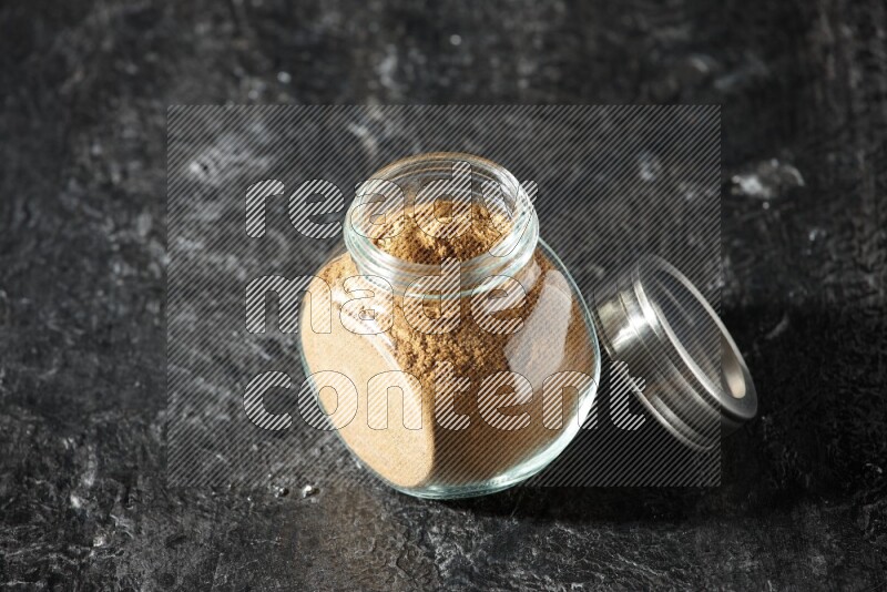 A glass spice jar full of allspice powder on a textured black flooring