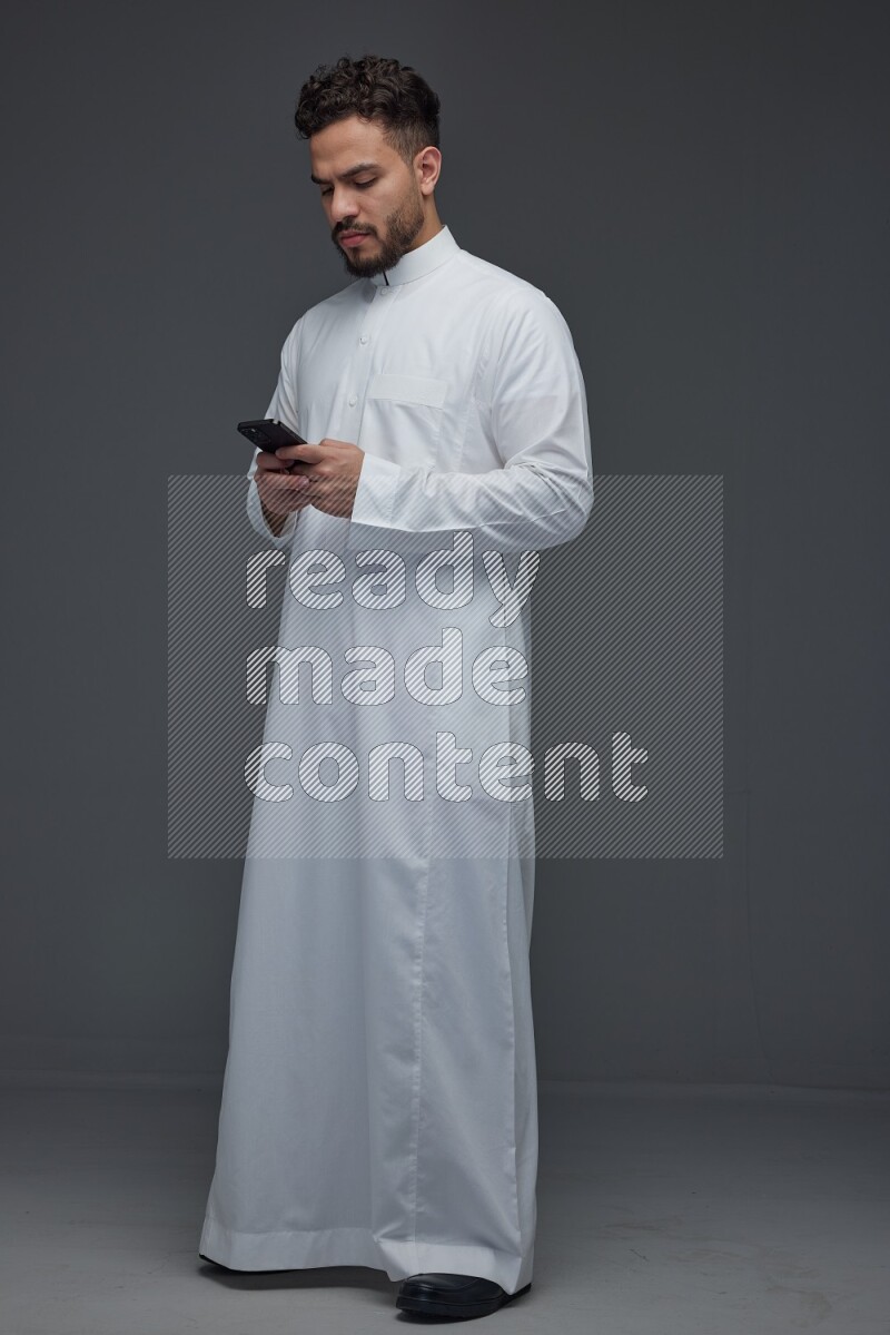 A Saudi man wearing Thobe standing and using his phone eye level on a gray background