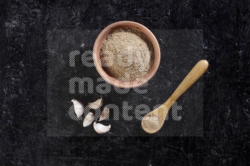 A wooden bowl and spoon full of garlic powder and beside it garlic cloves on a textured black flooring