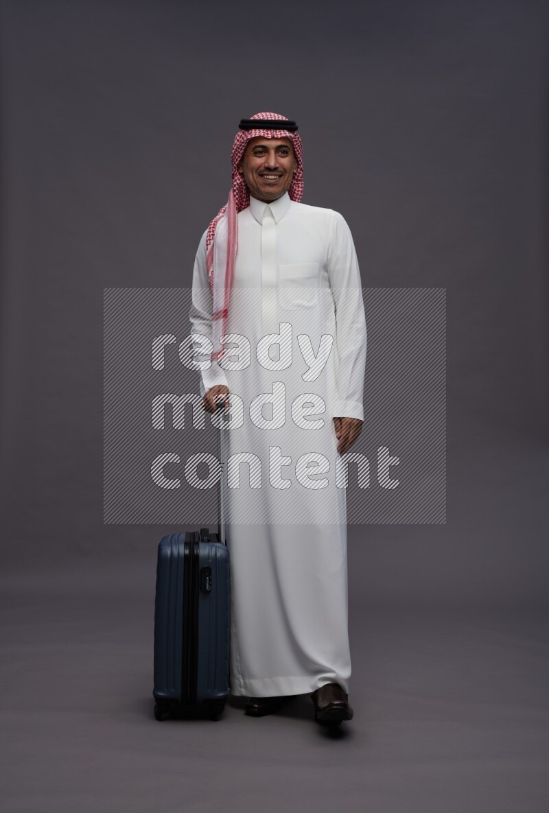 Saudi man wearing thob and shomag standing holding bag on gray background