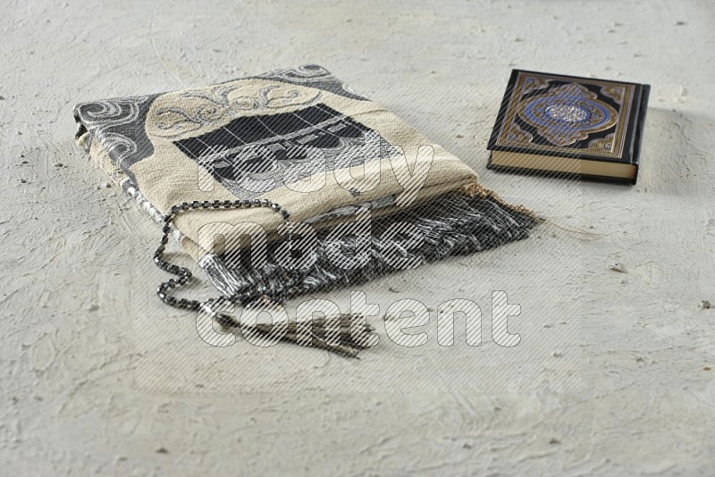 A folded prayer mat with different elements such as quran and prayer beads on white textured background