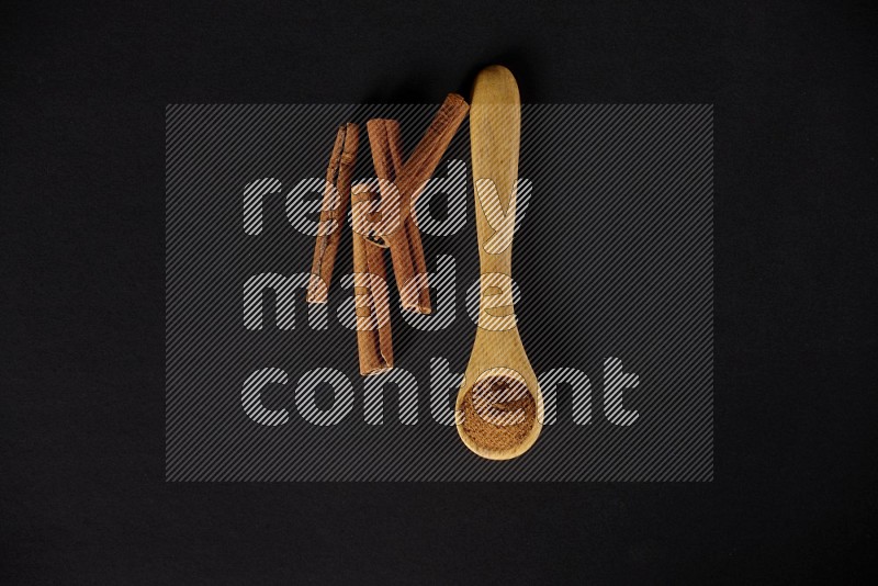 Cinnamon powder in a wooden spoon with cinnamon sticks beside it on black background