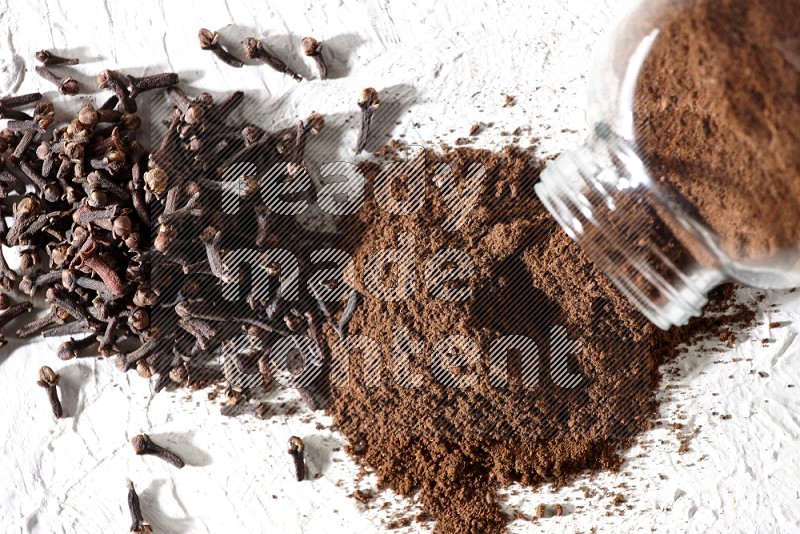 A glass spice jar full of cloves powder flipped and powder came out of it with cloves spread on textured white flooring