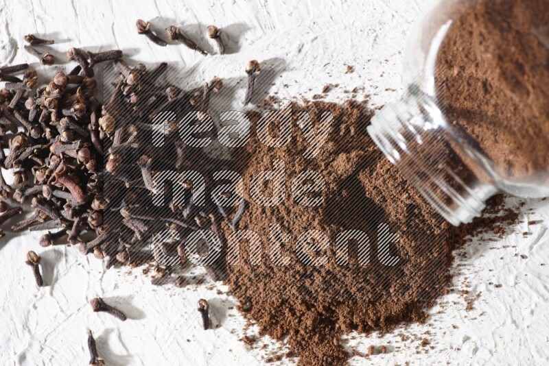 A glass spice jar full of cloves powder flipped and powder came out of it with cloves spread on textured white flooring