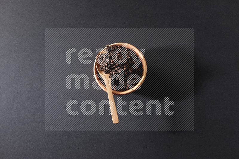 A wooden bowl and a wooden spoon full of cloves on a black flooring