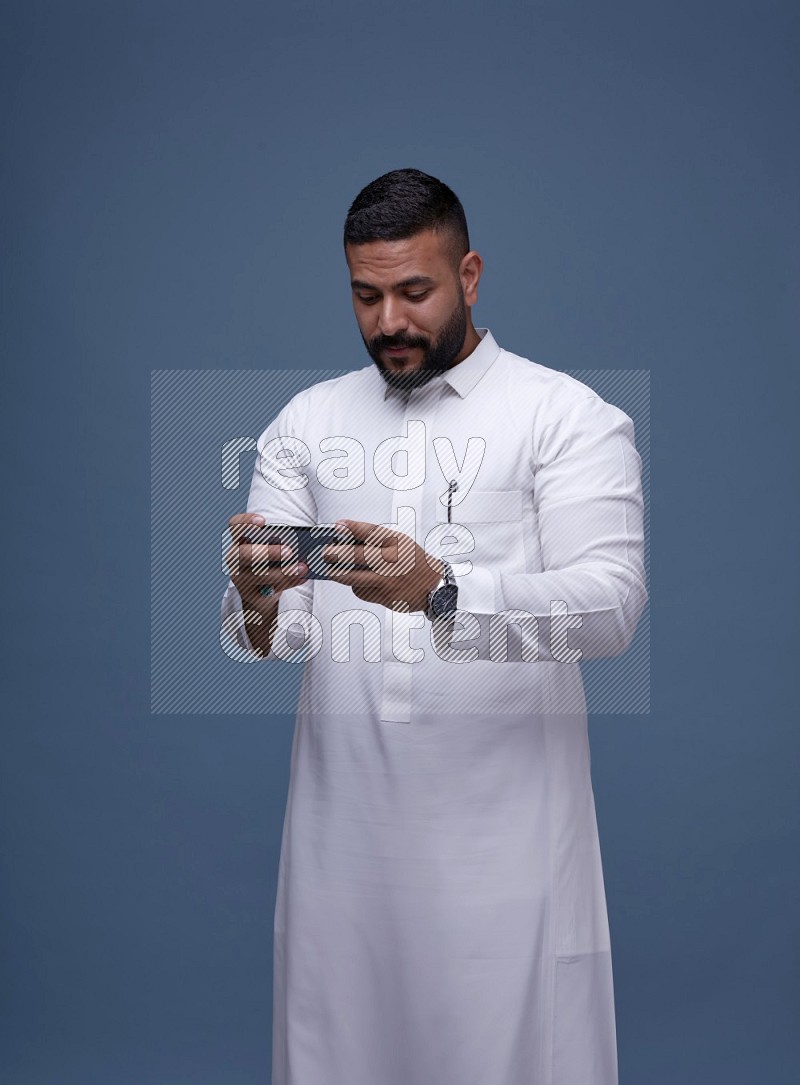 A man Playing a Game on his Smartphone  on Blue Background wearing Saudi Thob