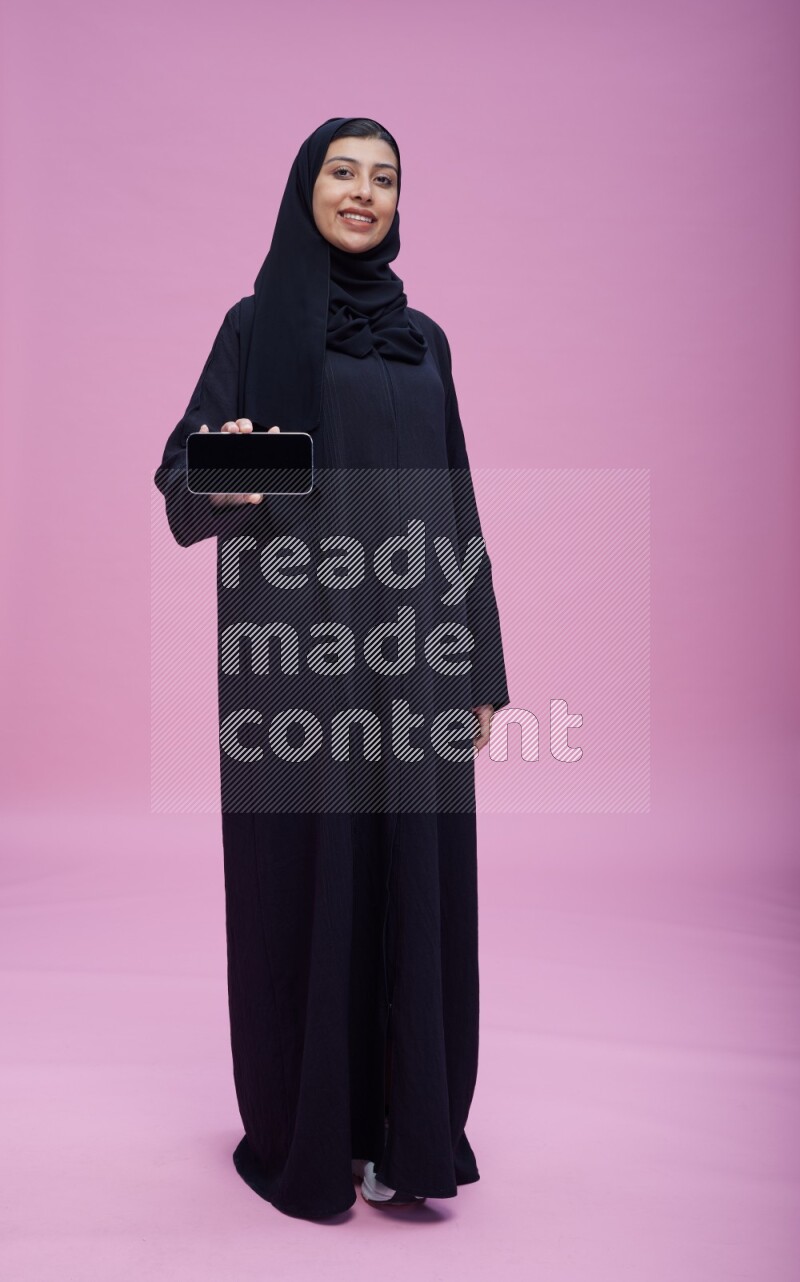 Saudi woman wearing Abaya standing showing phone to camera on pink background