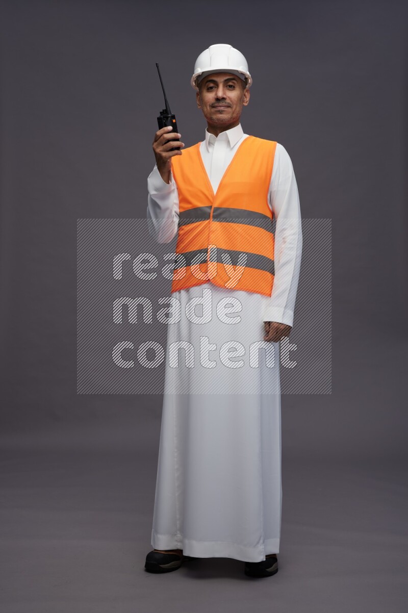 Saudi man wearing thob with engineer vest standing holding walkie-talkie on gray background