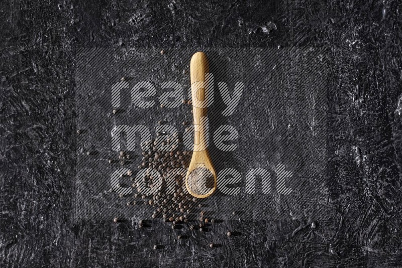 A wooden spoon full of black pepper powder and black pepper beads spread on a textured black flooring
