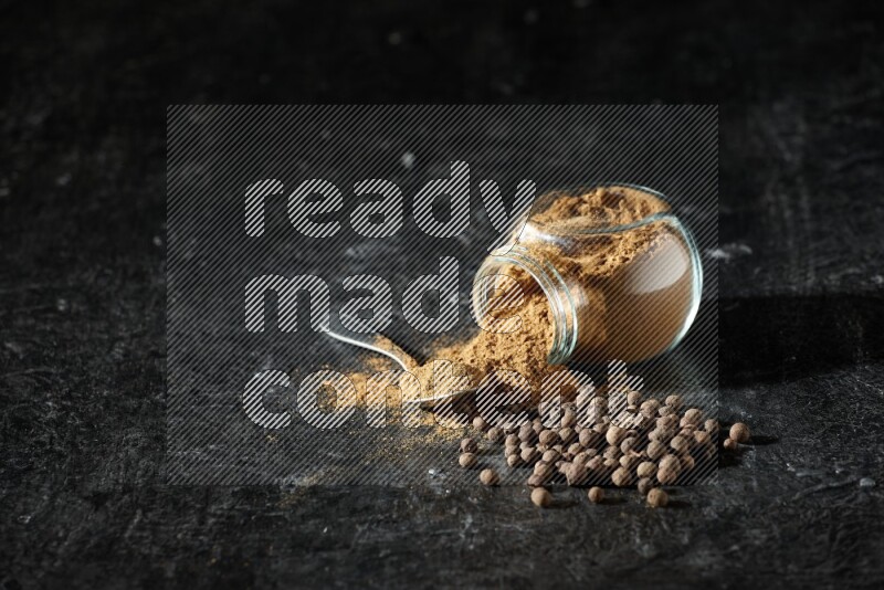A flipped glass spice jar and metal spoon full of allspice powder and powder spilled out of it with whole balls on a textured black flooring