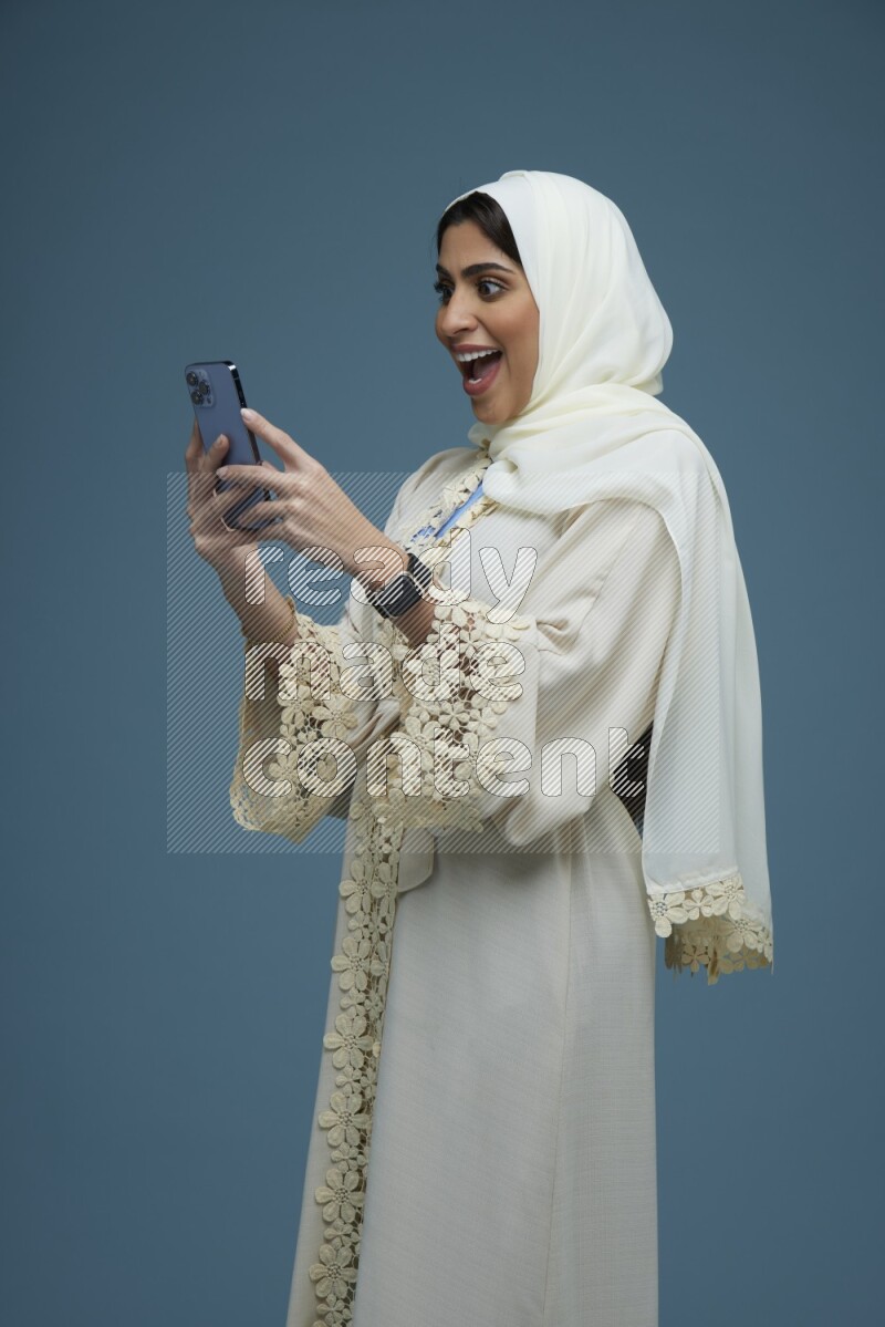 A Saudi woman Texting in a blue background wearing an off-white Abaya Hijab