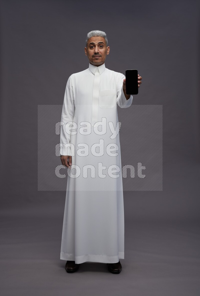 Saudi man wearing thob standing showing phone to camera on gray background