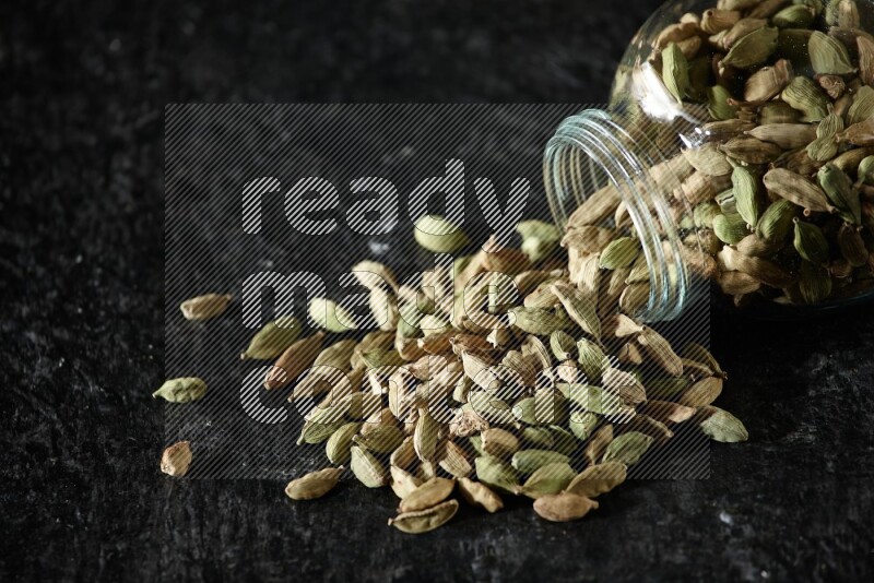 A flipped glass spice jar full of cardamom seeds on textured black flooring