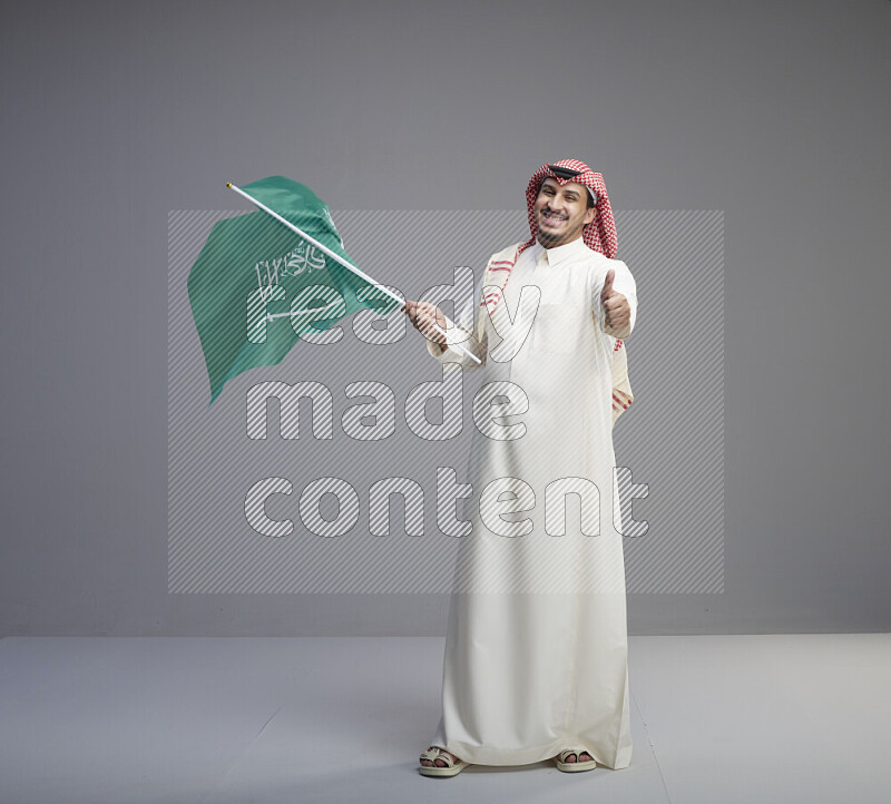 A Saudi man standing wearing thob and red shomag raising big Saudi flag on gray background