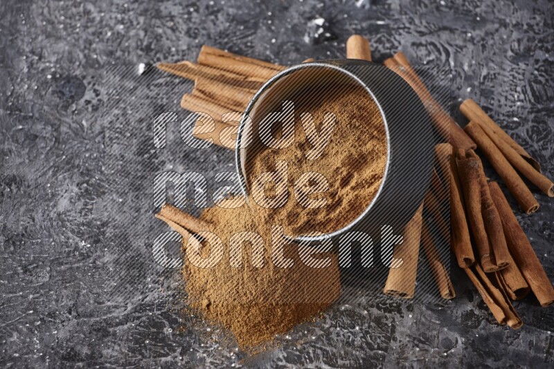 Black pottery bowl over filled with cinnamon powder and cinnamon sticks around the bowl on a textured black background