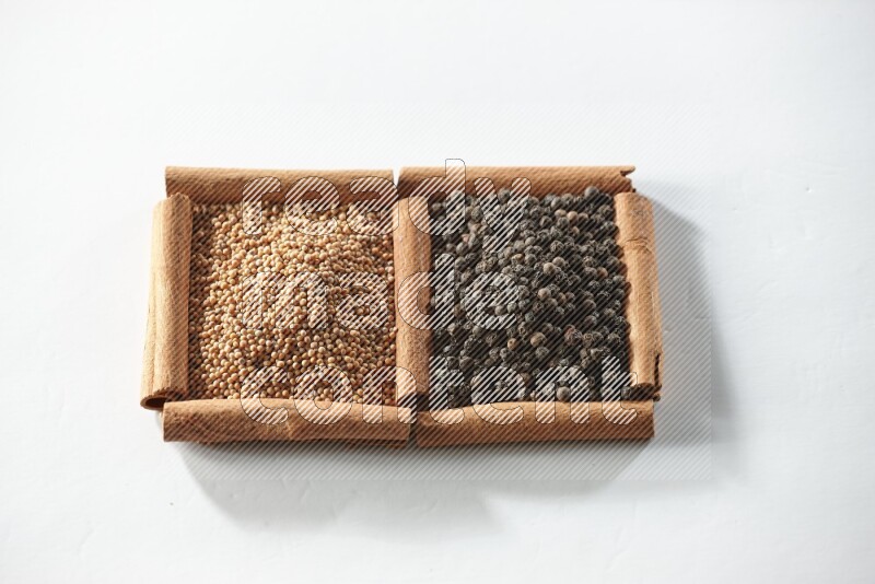 مربعان من أعواد القرفة ممتلئان بحبوب الخردل وحبوب الفلفل الأسود علي خلفية بيضاء