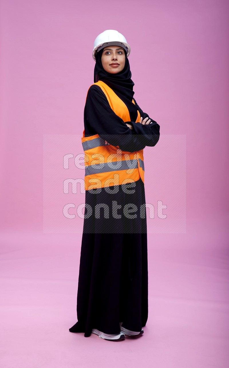 Saudi woman wearing Abaya with engineer vest and helmet standing with crossed arms on pink background