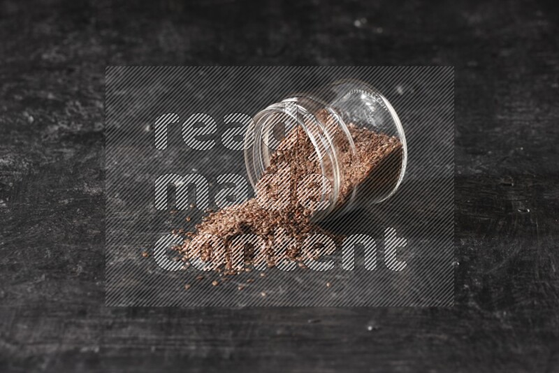A glass jar full of flaxseeds flipped and seeds spread out on a textured black flooring