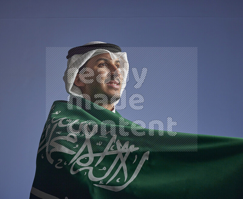 A close-up shot of Saudi man wearing thob and white shomag wrapping big Saudi flag on gray background