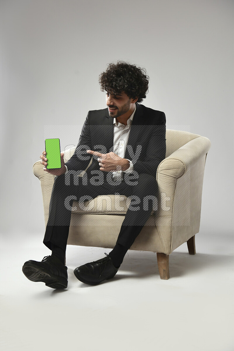 A man wearing formal sitting on a chair showing a phone screen on white background