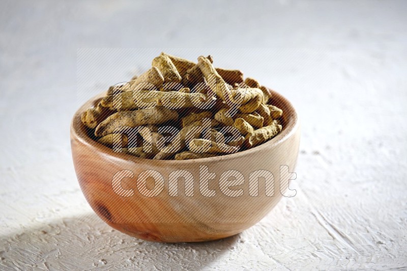 A wooden bowl full of dried turmeric whole fingers on a textured white flooring