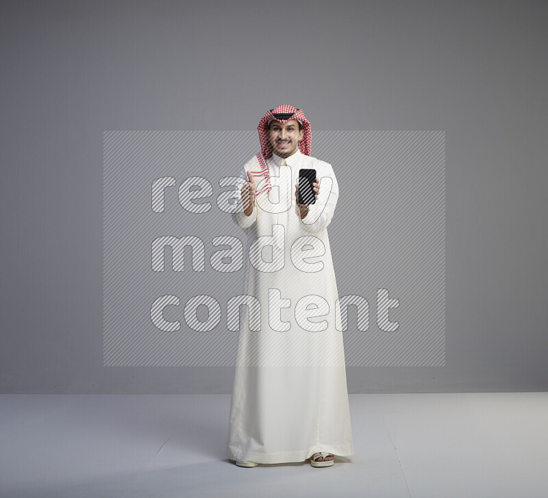 A Saudi man standing wearing thob and red shomag showing phone to camera on gray background