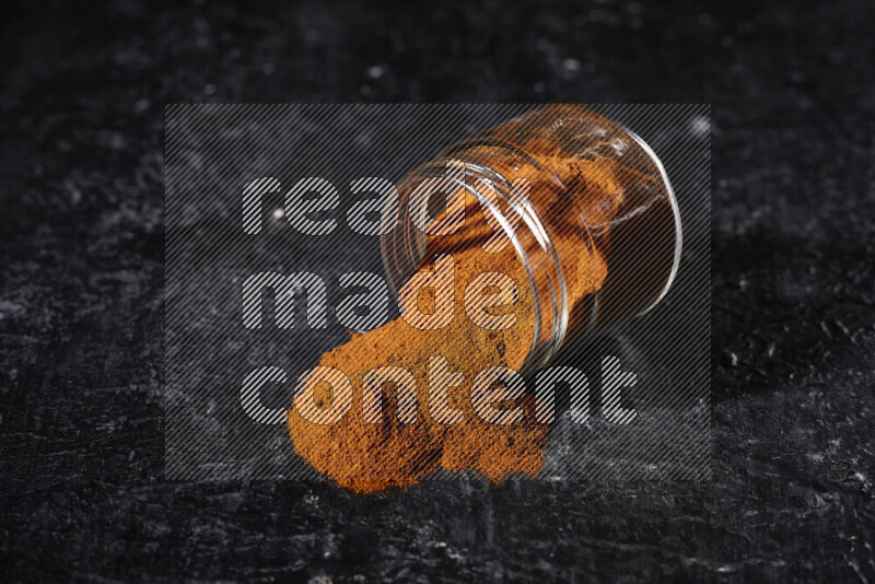 A glass jar full of ground paprika powder flipped with some spilling powder on black background