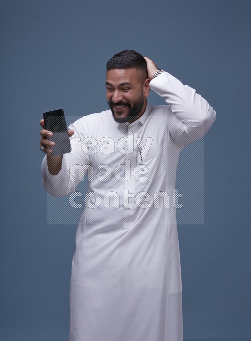 A man Showing his phone in Blue Background wearing Saudi Thob