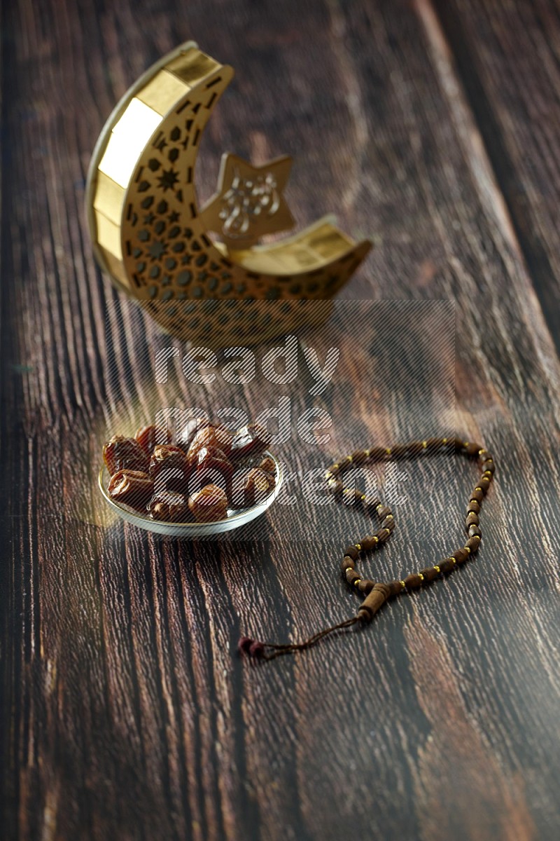 A wooden golden crescent lantern with different drinks, dates, nuts, prayer beads and quran on brown wooden background