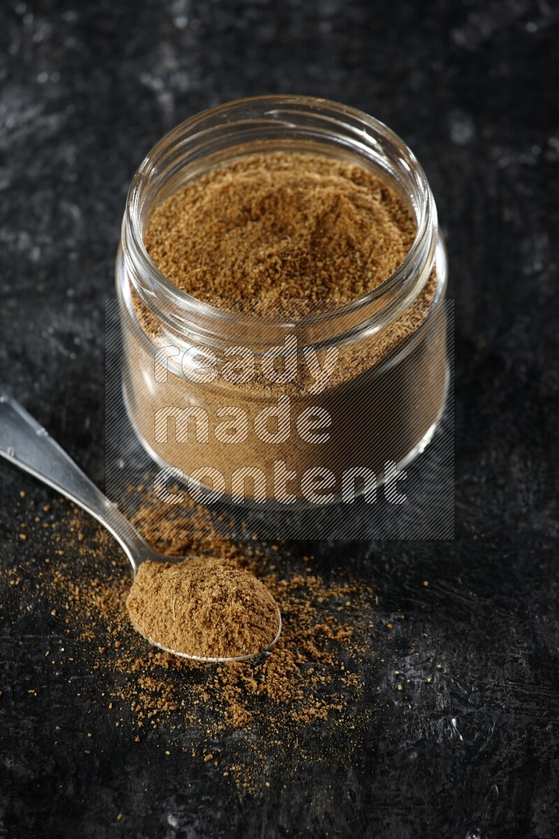 A glass jar and a metal spoon full of cumin powder on a textured black flooring