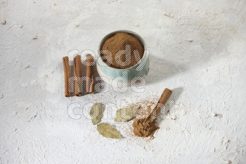 Cinnamon powder in a ceramic bowl with cinnamon sticks and laurel leaves on white background