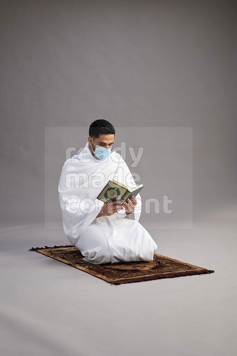 A man wearing Ehram with face mask sitting on floor reading quran on gray background