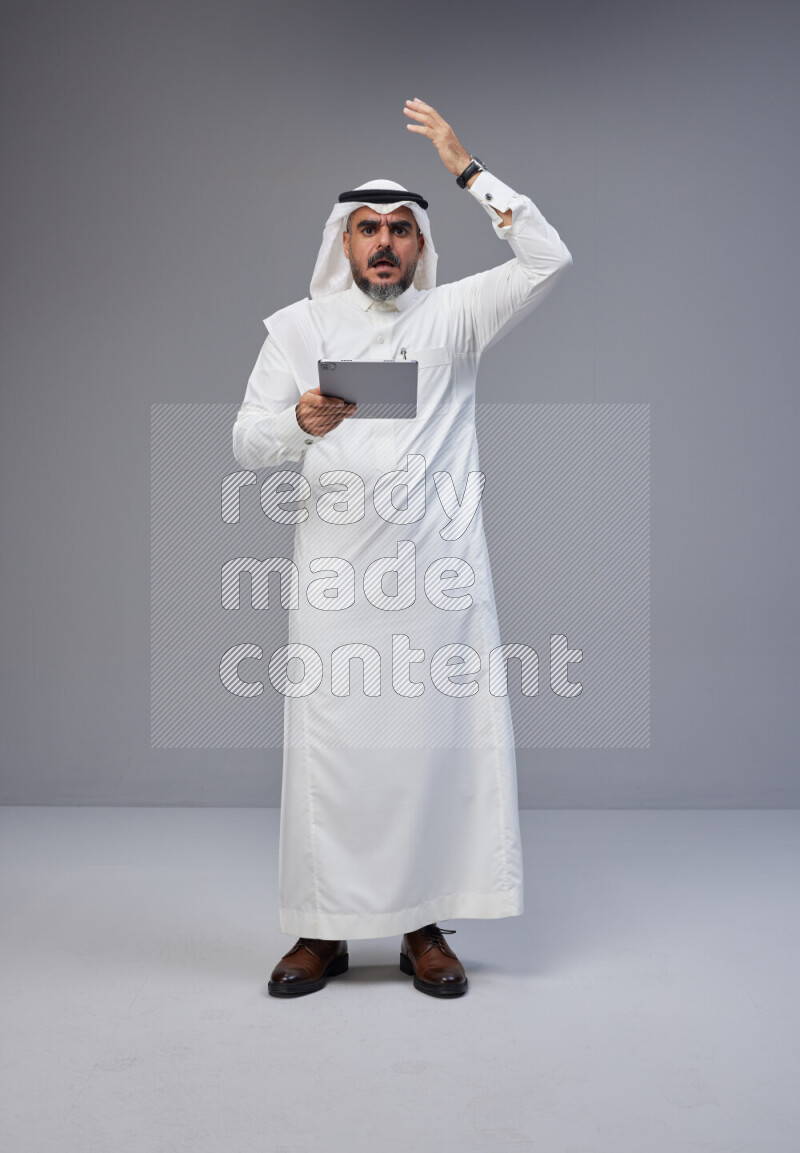 Saudi man Wearing Thob and white Shomag standing working on tablet on Gray background